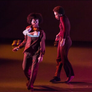 Two dancers walk on stage, one facing forward one facing stage right