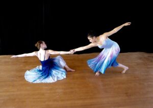 Two dancers in blue and white dresses share weight. One of them sits on the floor, pulling the other one closer.