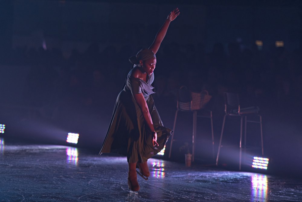 Dancer with ice skates swings arms to a vertical line, one arm reaches down the other reaches up as their upper body is hinged forward.
