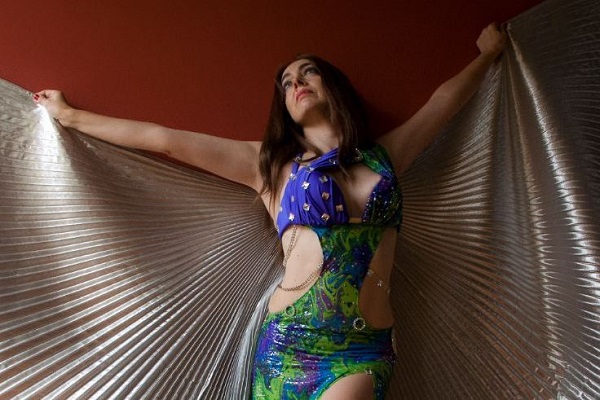 Dancer holds metalic fabric behind their back with arms open wide, revealing blue and green costume