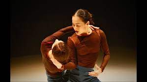 two dancers in brown longsleeve shirts partner. One holds the other by the neck in a hinged position.