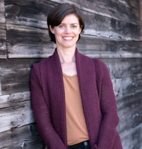 Christina, a pale skinned femme presenting person with short dark hair, stands agains a dark wooden wall and smiles at the camera.