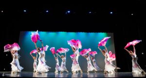 Company of dancers on stage wearing white long dresses reach up holding pink fabric and extend right leg forward.