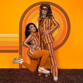 A person in roller skates kneels down while another stands with hands on hips. They both pose in front of a orange wall with a striped pattern.