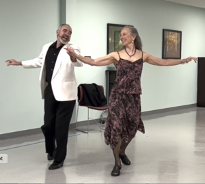 Two dancers hold hands sideways and reach opposite arm outwards as they step their outmost foot back.