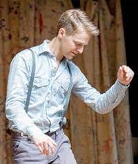 Nic Gareiss, wearing a button down shirt and pants with suspenders, looks down and raises left arm gently while dancing.