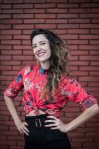 Mariel Gastiarena, a pale-skinned femme presenting dancer, wears a red shirt with flowers and rolled up sleeves and poses with hands on hips and smiles to the camera. Background is a brick wall.