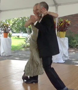 Two tango dancers, one in a long white dress and another in a black suit dance together, embraced, in an open space with wooden floors.
