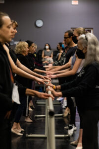 Multiple dancers of different ages, abilities, genders and ethnicities hold on to a ballet barre.