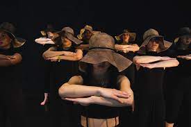 Dancers with hats covering most of their faces and wearing black costumes, stack their forearms in front of their own chests.