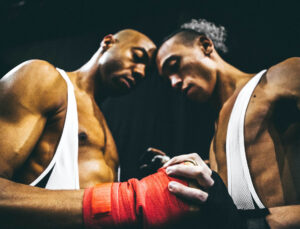 Two male-presenting dancers touch foreheads and wear white wrestling tank tops and hand wraps while one of them holds the other's fists.