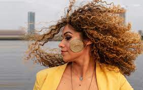 Woman-presenting person looks over right shoulder while her curly hair blows in the wind.