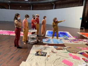 Six people stand in the Cyclorama space at BCA as one of them extends their arm to one of the areas of the wall in front of them and opens their hand. Large canvases are spread out on the floor with pink and blue paint on them.