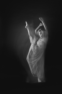 Black and white delayed image of dancer in a dress kicking one leg up and reaching both arms up softly.