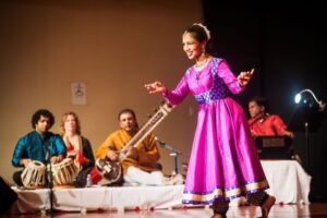 Shefali Jain on stage in a pink dress dances Kathak in front of a live group of musicians.