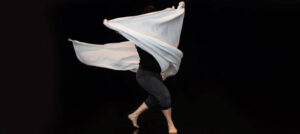 Dancer twirls with arms in the air holding a long white fabric in a way that the fabric covers the dancer's upper body.
