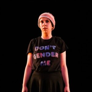 Dancer with beanie and shirt that reads"don't gender me" stands in a dark space.