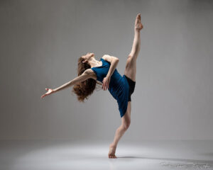 Ava Girard in blue dress kicks left leg up while hinged back on a forced arch, swinging arms back