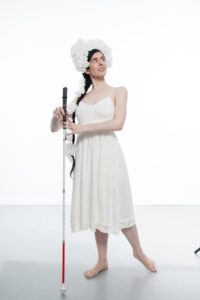 Krishna Washburn, from Movement Research, holds a guide cane and looks up towards her left. She wears a long white dress and had many white flowers on her head and along her long dark hair.