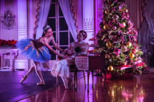 Two ballerinas wearing tutus and pointe shoes in a purple lit living room with a Christmas tree. One holds the nutcracker doll as the other stands and leans forwards reaching for the doll.