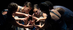 Five dancers gather under a spotlight and hold hands as if holding an imaginary sphere.