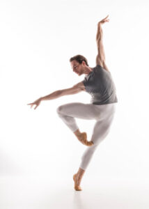 Male-presenting ballet dancer in white tights and grey tank top is on an off-balance rélevé passé reaching arms in an L shape.