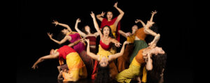 Ananya Dance Theatre dancers in vibrant earth tones reach are close together forming a circular shape and reach their arms outwards, most of them in back bends and the center dancer looks straight at the camera.