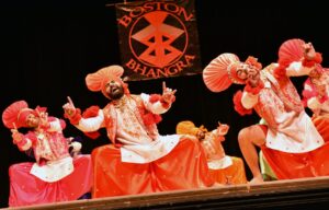 Bhangra Dancers on stage in a deep bend of both knees while legs are wide and reaching arms forward.