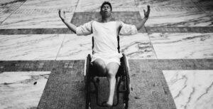 Black and white photo of a dancer in a wheel chair reaching rounded arms out to the sides and looking up.
