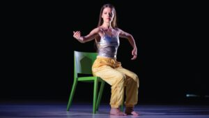 Dancer sits on green chair and holds arms up in the air as if holding an imaginary guitar