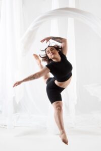 Maria leaps with right leg straight and left leg bent behind them; left arm bent over head and right arm straight to the side. Maria wears a black cropped shirt and black spandex shorts and is in a white space with white fabric hanging all around.