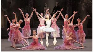 Corps de ballet in pink tutu's surround leading ballerina in a white tutu who stands on sous-sous in the center of the circle with arms up in a V position.