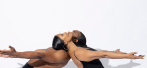 Two black dancers rest their heads on each other's shoulders leaning back and looking up while reaching arms forward.