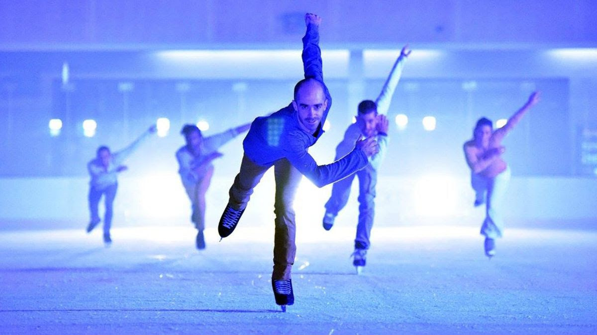 Five ice-skaters skate towards the camera leaning forward.