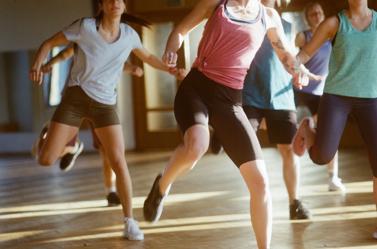Multiple people in a wooden floor space exercising, lifting one leg.