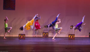 Six dancers on stage: three handstand on three separate boxes and three hop with one leg bent.