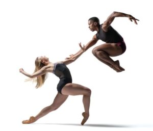 Two BalletX dancers: one on pointe with long blonde hair lunges over right leg and reaches arm towards the other dancer who jumps with both legs bent towards chest, reaching downward at blonde dancer and rounding other arm behind upperbody.