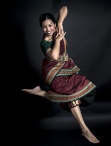 Smitha smiles and jumps with one leg bent and the other straight while fingers are interlaces and elbows are tilted towards the camera.