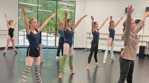 Young dancers in leotards, tights and warm ups reach arms up and extend their legs on the tips of their toes making an X shape with their bodies.
