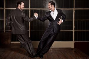 The tango twins in black suits hold hands and lean back away from each other.
