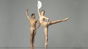 Two ballet dancers in nude tight briefs and top: one stands on tip toes with legs zipped, one arm rounded up and one hand on hip; the other on pointe in an arabesque holding up a fan overhead and with other hand on hip.