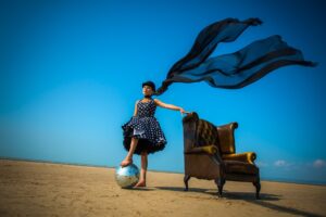 Chiara D'Anna in a polka dot dress and scarf blowing in the wind, standing in a dessert-like space holding on to a brown arm chair and placiing one foot over a metalic reflective sphere.