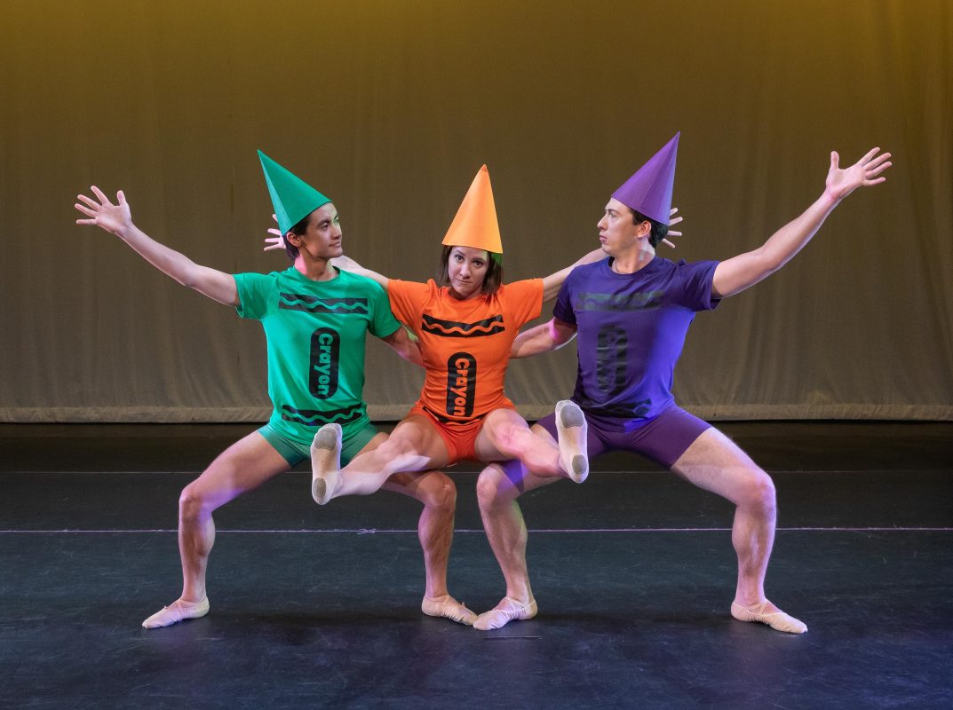 Three dancers dressed as crayons on stage. Two, side by side, squat down with third in the middle sitting on their thighs.