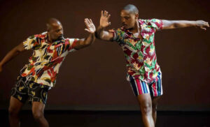 Two dancers in colorful patterned shorts and short-sleeve button down shirts look at each other while pressing forearms against each other's and reaching their outside arm outward.