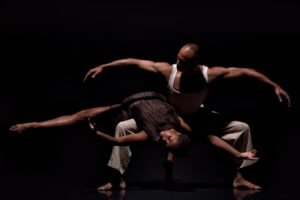 Two dancers in a dim lit stage, one lays back on the other's leg as the other bends both legs deeply and rounds arms wide.