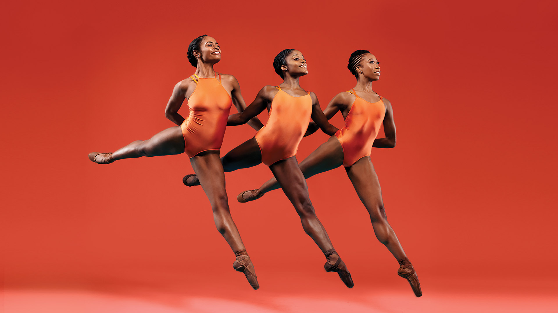Three dancers in pointe shoes and orange leotards jump with their left legs straight down and their right legs straight back while interlacing arms and holding hands behind their backs.