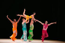 Paul Taylor Dance Company on stage wearing different solid bright colored costumes. Four of them travel stage left while holding on to a fifth dancer over head who bends one leg in the air and faces down.
