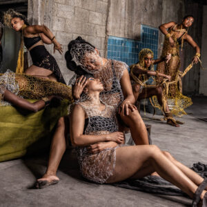 Photo of Urban Bush Women in metalic, chain-like costumes i different poses. In the forefront are two company members: one sits on the floor and lays back on the others thigh and chest as the latter embraces the first's head gently