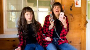 Photo of two performers in red plaid shirts with black shirts underneath and jeans, with the same bangs and hair cut, one resembling a mom, and the other, a daughter. Both sit on a bench outside a house and the mother-figure holds on to a landline phone and a smartphone in despair while the daughter-figure seems to be confused.