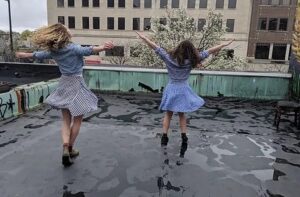 Two dancers in checkered dresses twirl facing away from the camera with arms up on a rained-on rooftop.